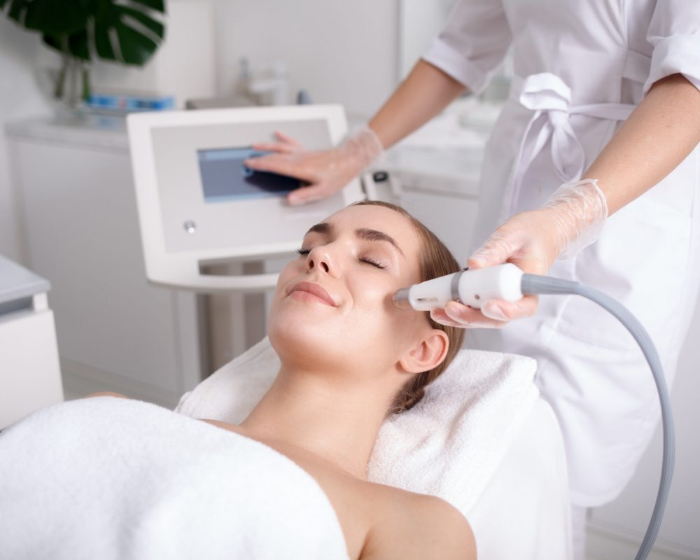 Side view of happy young woman getting cavitation rejuvenating skin treatment at spa. She is lying on massage table and smiling. Beautician is touching monitor screen while holding tool near female cheek